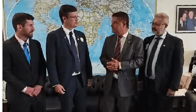 Quatro homens em trajes formais conversando em um ambiente com um mapa-múndi ao fundo.