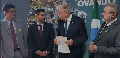 Quatro homens estão em um ambiente formal, com uma bandeira ao fundo, discutindo um documento importante.