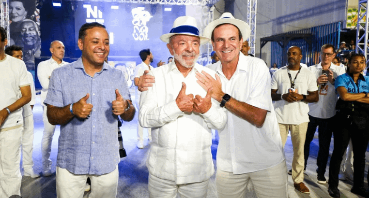 Três homens sorrindo e fazendo gestos de positivo, vestindo roupas brancas. O fundo apresenta uma decoração festiva com luzes e pessoas ao redor.