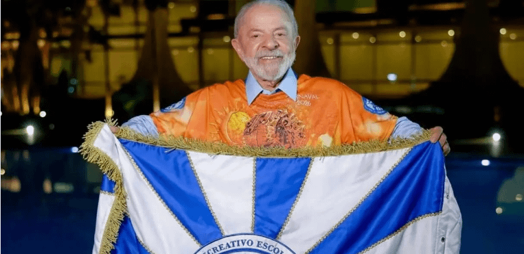 Homem sorridente segurando uma bandeira branca e azul, vestido com uma camiseta colorida de Carnaval, em frente a um ambiente iluminado.
