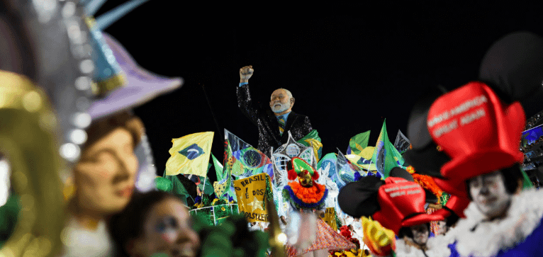 Um homem vestido de forma elegante, levantando o punho em celebração, destaca-se em um ambiente festivo do Carnaval, cercado por pessoas em fantasias coloridas e bandeiras do Brasil.