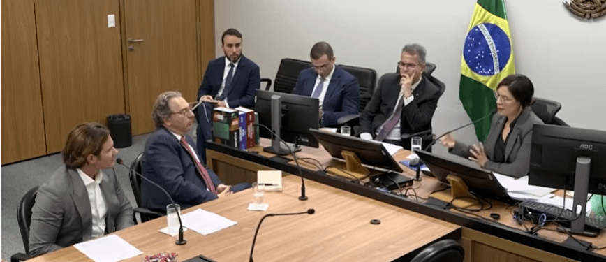 Audiência com vários homens e uma mulher, discutindo temas de interesse jurídico, com a bandeira do Brasil ao fundo.